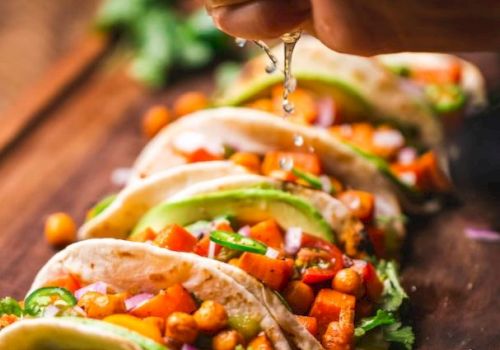 The image shows five tacos filled with vegetables and chickpeas, garnished with cilantro and lime slices. A hand is squeezing lime juice over them.