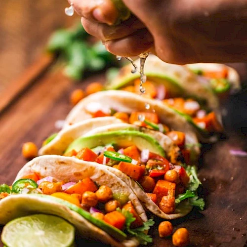 The image shows five tacos filled with vegetables and chickpeas, garnished with cilantro and lime slices. A hand is squeezing lime juice over them.