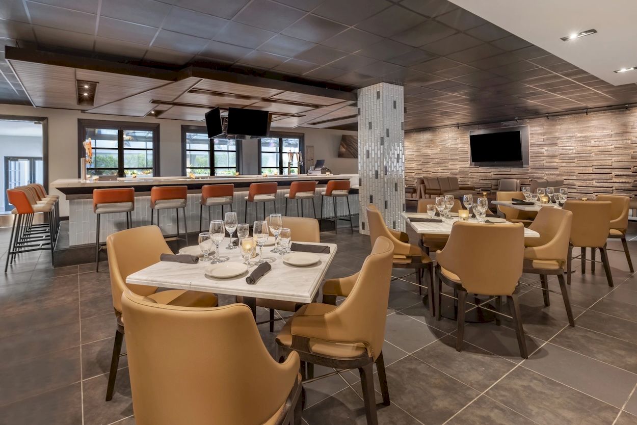 A modern banquet room with set tables, wine glasses, bar stools, and TVs on brick walls.
