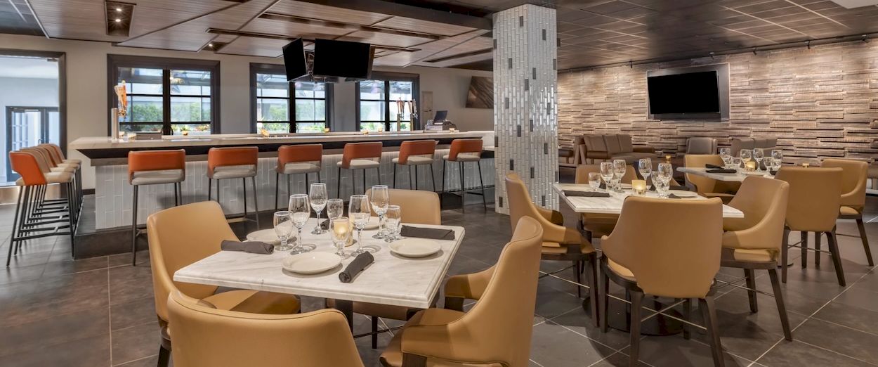 A modern banquet room with set tables, wine glasses, bar stools, and TVs on brick walls.