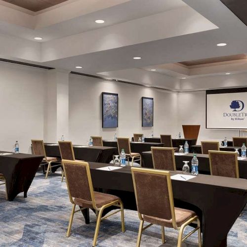 The image depicts a conference room with tables, chairs, water bottles, artwork on the walls, and a projection screen displaying a DoubleTree logo.