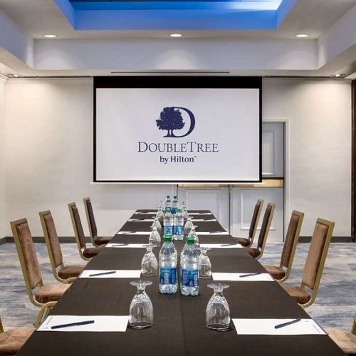 The image shows a conference room setup with a table, chairs, water bottles, and papers. There is a screen displaying 