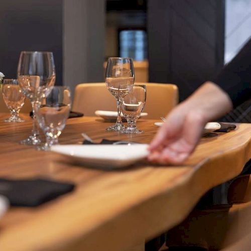 A person arranging a place setting in a modern, elegantly set dining area with wine glasses, napkins, and a floral centerpiece ends the sentence.