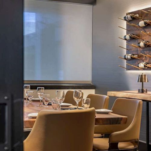 A cozy dining area with a wooden table, beige chairs, wine glasses, and wine bottles displayed on the wall, featuring a modern and elegant décor.