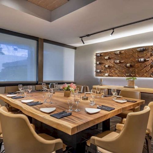 A modern dining room with a wooden table set for a meal, beige chairs, wine bottles displayed on the wall, and minimalistic decor.