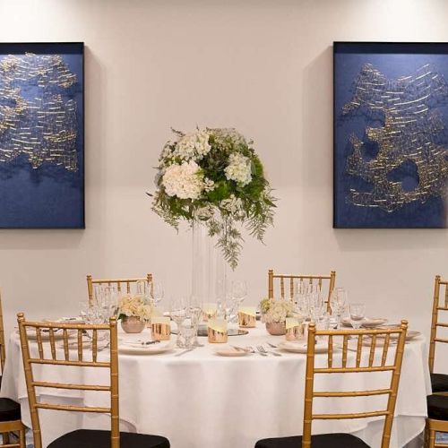 A round table set for an event, adorned with a floral centerpiece and surrounded by six chairs, with two abstract wall arts in the background.