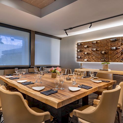 A stylish dining room with a wooden table set for eight, modern chairs, wine shelving, and elegant tableware, under soft lighting.