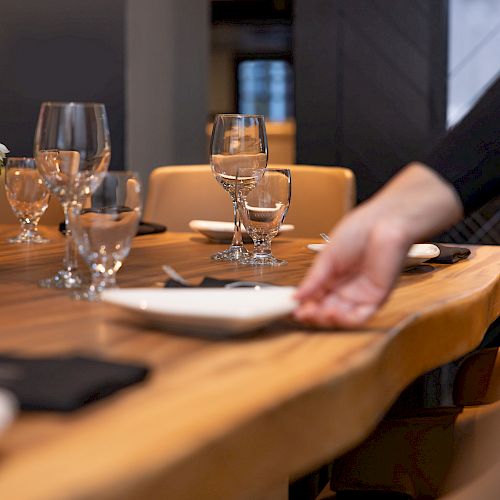 A hand is setting a dining table with plates, glasses, and napkins. There's a centerpiece with flowers on the wooden table.