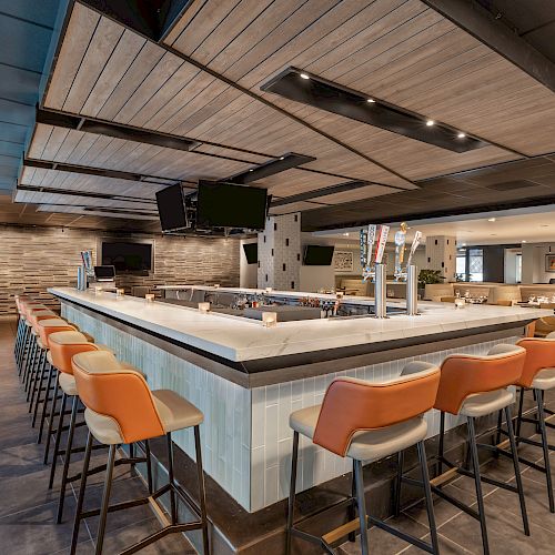 A modern bar with a rectangular counter, orange-cushioned barstools, several taps, and multiple TV screens mounted on the ceiling.