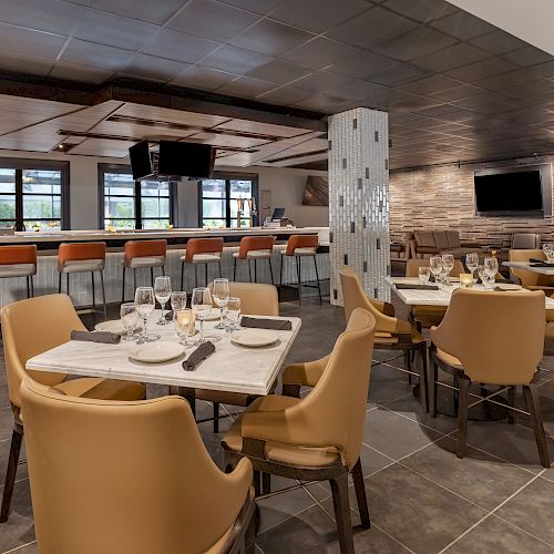 Nice restaurant interior with a modern design, featuring tables set for dining, brown chairs, a bar with orange stools, and a TV on the wall.