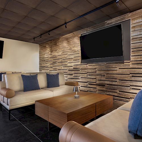 A modern lounge area with beige couches, dark blue pillows, a wooden coffee table, and two large TVs mounted on a textured wall with a potted plant in the corner.