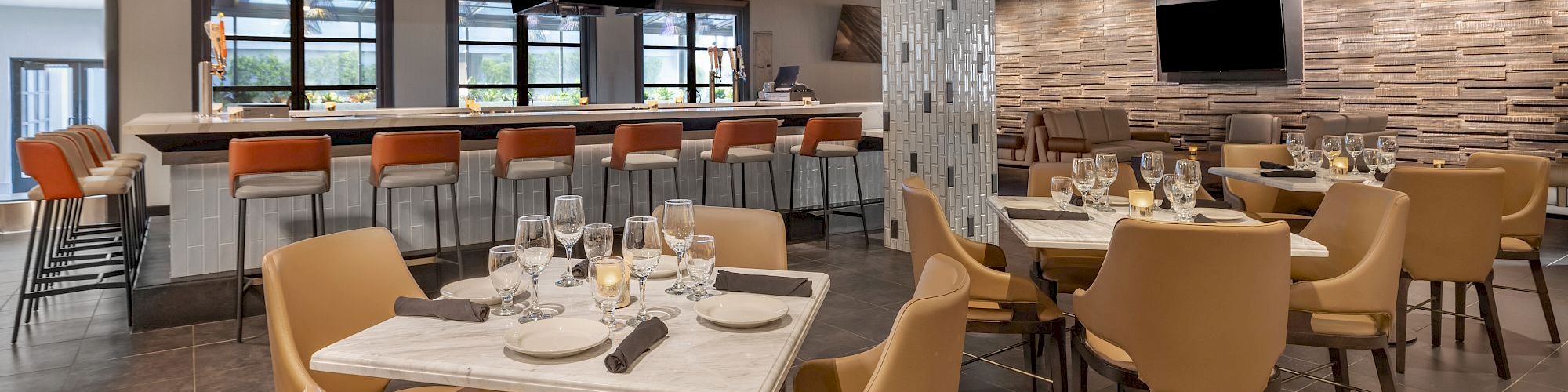 A modern restaurant interior with beige and orange seating, marble tables, set dining tables, and a bar area with high stools and a TV on the wall.