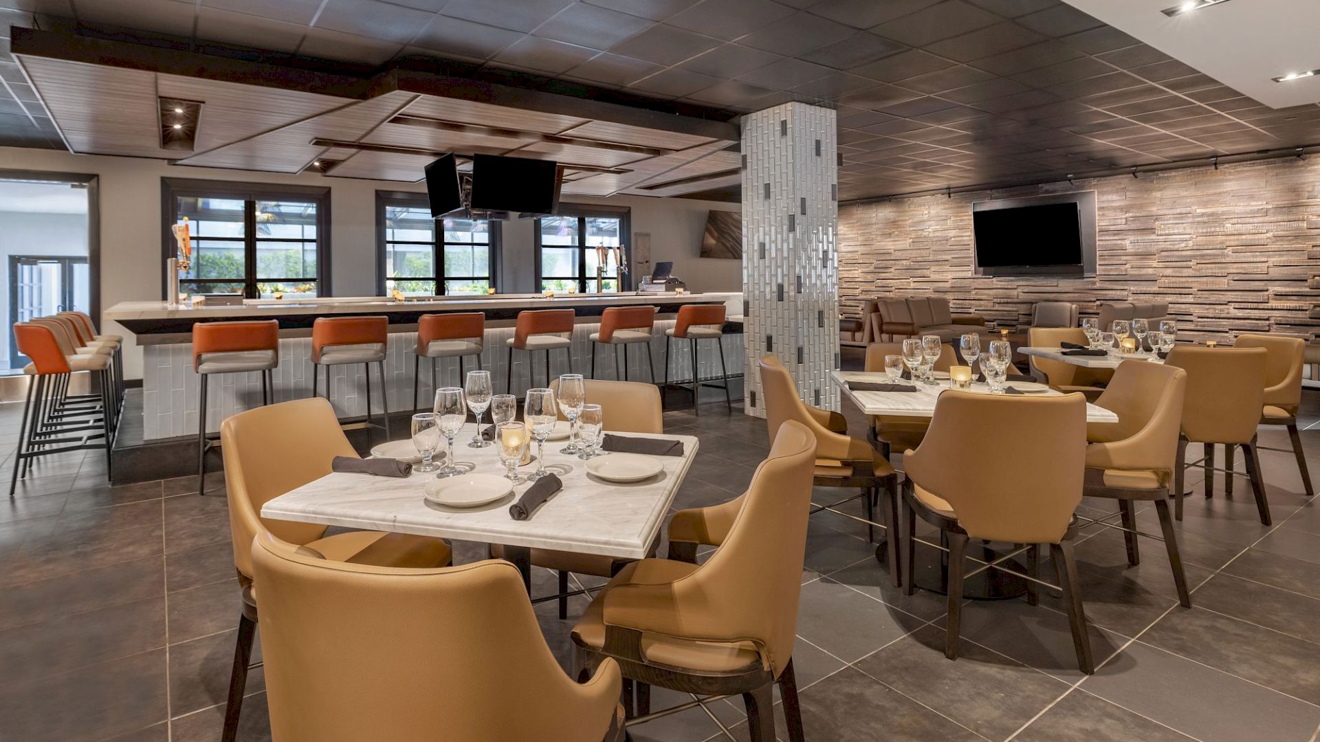 A modern restaurant interior with a bar area, orange chairs, beige dining chairs, set tables, and a TV on the wall.