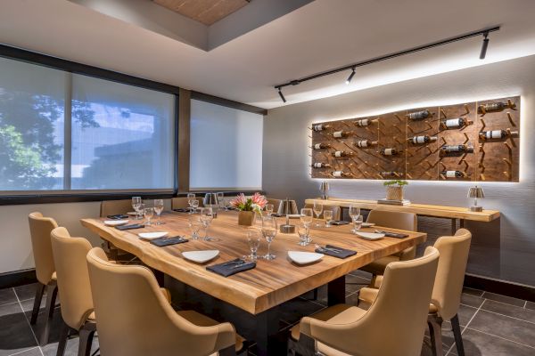 A modern dining room with a wooden table set for ten, wine bottles displayed on the wall, and large windows with shades drawn.