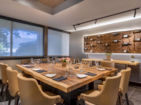 A modern dining room with a wooden table set for ten, wine bottles displayed on the wall, and large windows with shades drawn.