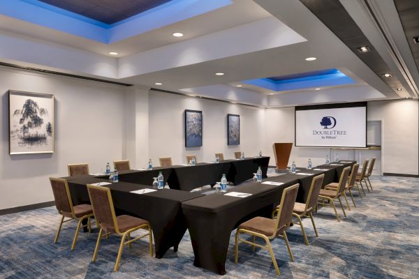 A conference room with a U-shaped table setup, water bottles, notepads, modern decor, and a screen displaying the DoubleTree by Hilton logo.