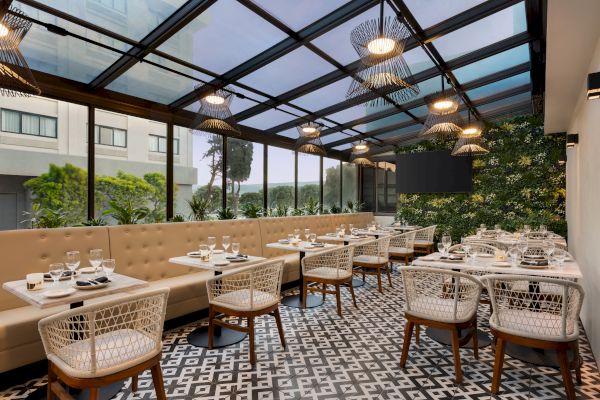 A modern, elegant restaurant with a glass ceiling, patterned floor tiles, cushioned bench seating, wicker chairs, and lush greenery along one wall.