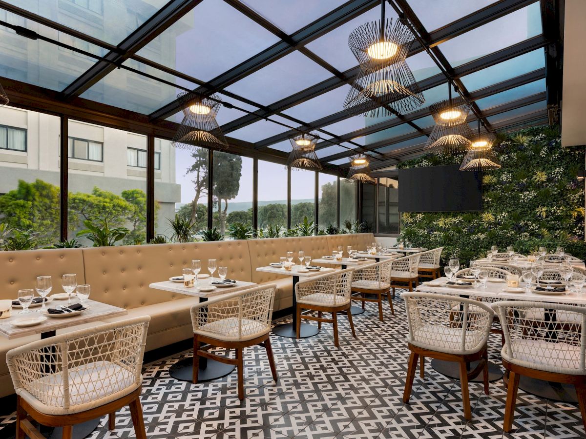 This image shows a modern restaurant with a glass roof, beige seating booths, and patterned floor tiles, set in a bright and lush environment, ending the sentence.