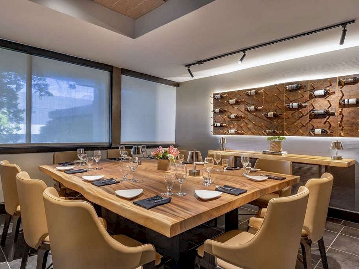 A modern restaurant dining room with a long wooden table set with place settings, chairs, and a wall wine-bottle display.