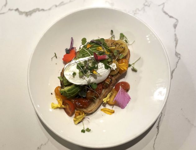 A plated dish with toasted bread topped with greens, poached egg, and colorful vegetables on a white marble surface.