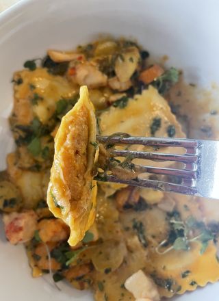 The image shows a close-up of ravioli, held by a fork, over a bowl containing more ravioli mixed with herbs and chunks of other ingredients.