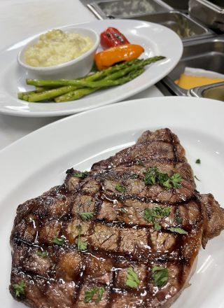 The image shows a grilled steak garnished with herbs on a white plate. In the background, there are mashed potatoes, grilled vegetables, and asparagus.