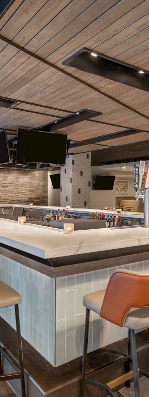 The image shows a modern bar with a square counter surrounded by barstools, overhead lights, and multiple TVs mounted on the ceiling.