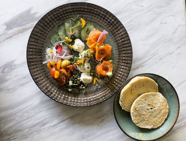 A beautifully plated dish with smoked salmon, various vegetables, and herbs in a textured bowl alongside a plate with two toasted bread slices.