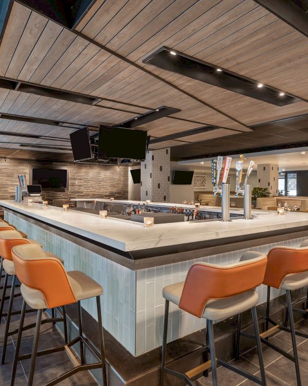 The image shows a contemporary bar with an L-shaped counter, orange chairs, several TVs, and modern lighting. The space is bright and clean.