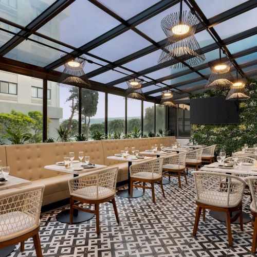 A modern restaurant with a glass roof, patterned floor, beige seating, and white wicker chairs is elegantly set and surrounded by greenery.