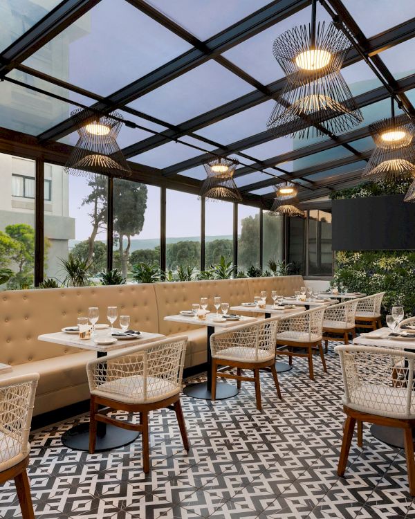 A modern covered patio dining area with beige banquettes, wicker chairs, patterned floor, and glowing wire pendant lights.