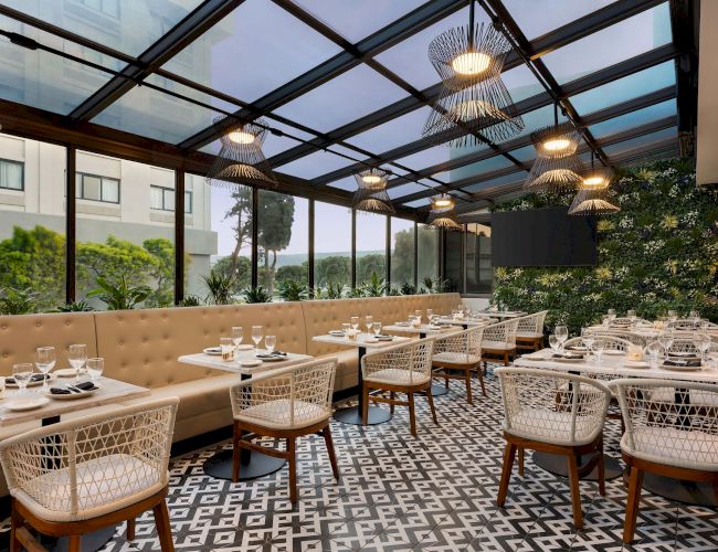 A modern restaurant with a glass roof, patterned floor, beige seating, and white wicker chairs is elegantly set and surrounded by greenery.