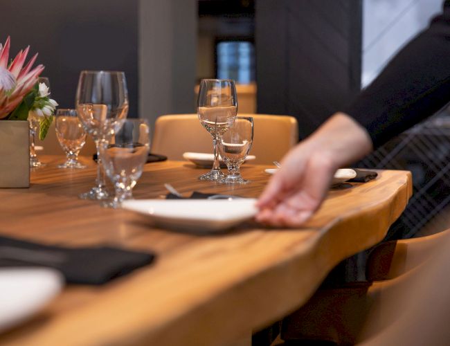 A hand is setting a dining table with plates, glasses, and napkins. There's a centerpiece with flowers on the wooden table.