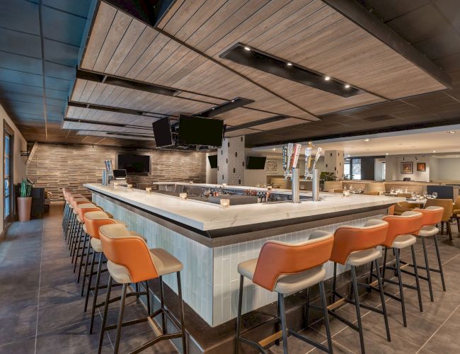 A modern event bar with a long white counter, orange stools, TVs, and a sleek wood-and-tile ceiling, lit by ceiling lights.