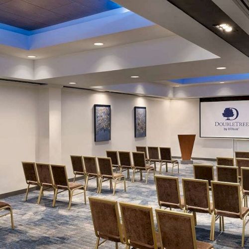 A modern meeting room with rows of chairs, framed art, and a projection screen reading DoubleTree by Hilton.