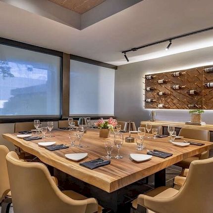 A modern restaurant or event room with a set wooden dining table, beige chairs, projector screen, and a wall display of bottles.