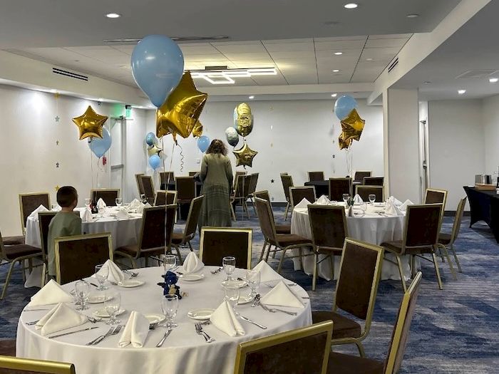 A set-up banquet room with round tables, white linens, cutlery, and blue/gold balloons (including star balloons) hanging overhead.